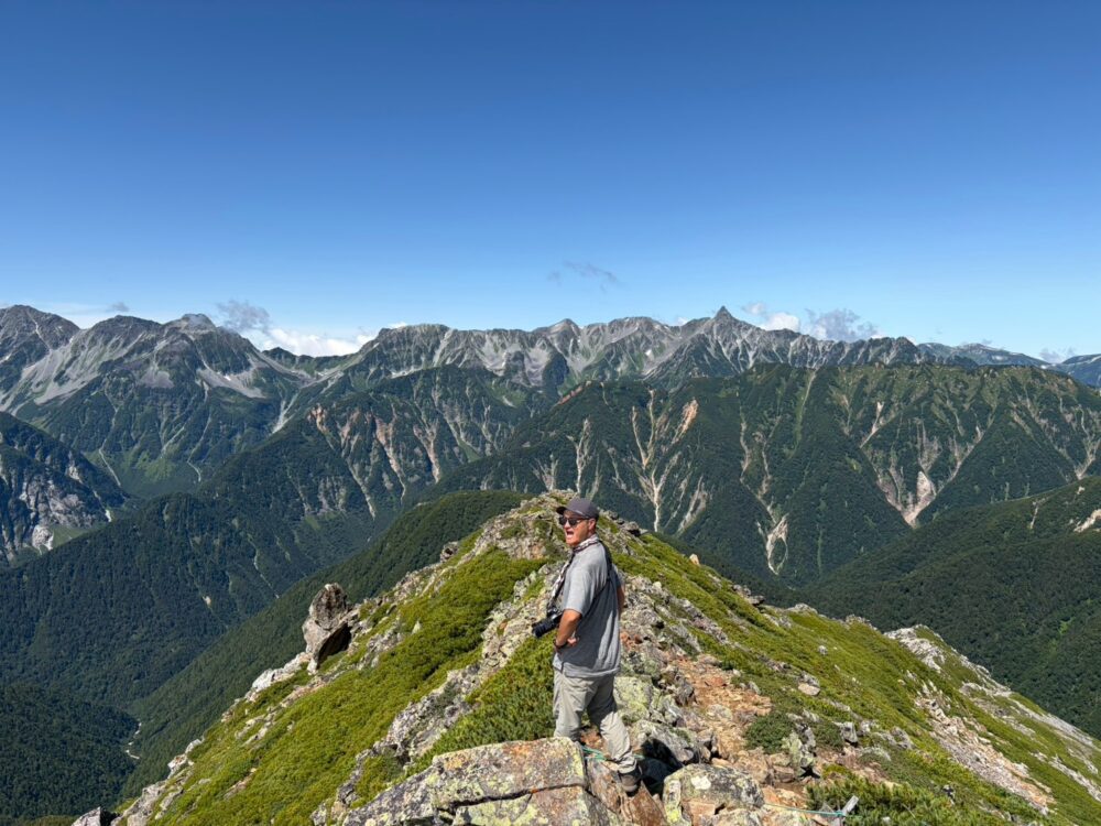 常念岳山頂で穂高連峰と槍ヶ岳をバックに写真を撮る登山者
